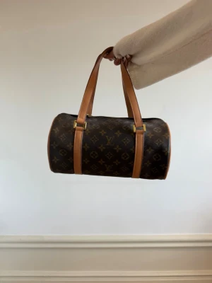 Louis Vuitton Monogram Papillon väska -  Louis Vuitton väska i klassiskt monogrammönster. Väskan har en rundad, tubformad design med bruna läderhandtag och detaljer i guld. Materialet är canvas med läderdetaljer. 