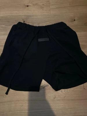 Svarta shorts från Essentials - Säljer ett par nästan helt nya svarta shorts från Essentials med elastisk midja och snörning. Shortsen har en enkel och clean design med fickor på sidorna och en diskret logga framtill. Perfekta för chill dagar eller när du vill ha en avslappnad stil.