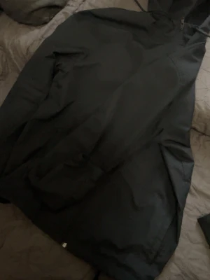 Svart vindjacka från Under Armour - Snygg svart vindjacka från Under Armour med dragkedja framtill och huva med dragsko. Jackan har en vit logga på bröstet och är tillverkad i ett lätt och tåligt material som passar perfekt när det blåser ute.
