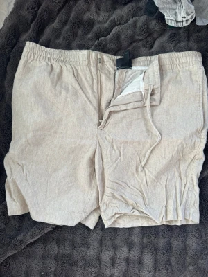 Beiga linneshorts från H&M - Snygga beiga shorts från H&M i regular fit med dragsko i midjan och gylf med knapp. Tillverkade i ett luftigt linnematerial som passar perfekt till varma dagar. Enkel och stilren design som är lätt att matcha med olika outfits.