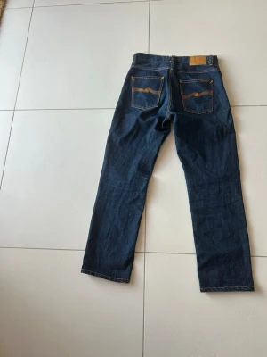 Mörkblå jeans från Nudie Jeans - Säljer ett par klassiska mörkblå jeans från Nudie Jeans med raka ben och kontrastsömmar. Jeansen har fem fickor och Nudie Jeans ikoniska broderi på bakfickorna. Tillverkade i slitstark denim, perfekta för en avslappnad och stilren look.