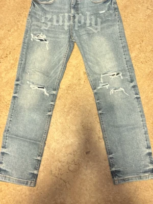 Ljusblå slitna jeans med tryck - Säljer ett par ljusblå jeans med raka ben och slitningar på både fram- och baksida. Jeansen har trycket 'supply' framtill och klassiska fem fickor. Perfekta för en avslappnad streetwear-look.