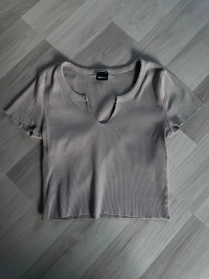 Beige ribbad topp från Gina Tricot - Snygg beige ribbad topp från Gina Tricot med korta ärmar och unik v-ringad halsringning. Croppad modell som är perfekt till jeans eller kjol. Enkel och stilren design som passar till många olika looks.
