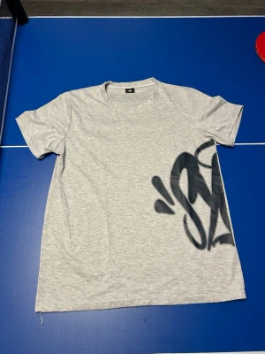 Grå t-shirt med svart print - Snygg grå t-shirt från 0% FUC*ERIES i storlek M. T-shirten har ett stort svart grafiskt tryck på ena sidan och är tillverkad i mjuk bomull. Perfekt för en avslappnad och trendig stil.