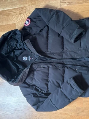 Canada Goose McMillan  - Säljer en svart dunjacka från Canada Goose med huva och klassisk logotyp på ärmen. Jackan har dragkedja och tryckknappar framtill samt två nonchalanta sidofickor. Perfekt för kalla vinterdagar och riktigt varm tack vare dunfyllningen.