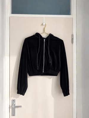 Svart croppad hoodie i velour - Snygg svart croppad hoodie i mjuk velour med dragkedja framtill och justerbar huva. Långa ärmar med resår vid ärmslut och midja. Perfekt för en chill och trendig look.