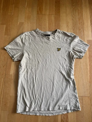 Beige / grå / grön  t-shirt från Lyle & Scott - Klassisk  t-shirt från Lyle & Scott med rund hals och korta ärmar. Färgen är beige/grön/grå. På bröstet sitter märket. 