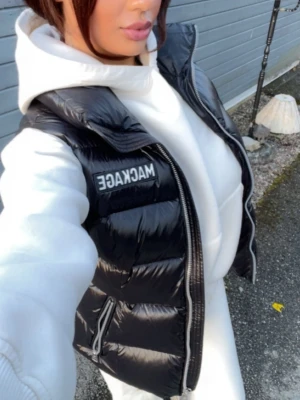 Svart dunväst från Mackage - Snygg svart dunväst från Mackage med hög krage och två dragkedjefickor. Västen har en glansig finish och tydliga quiltade partier. Märkeslogga på bröstet. Perfekt att slänga över en hoodie för en cool och sportig look. Använd ca 2/3 gånger! 
