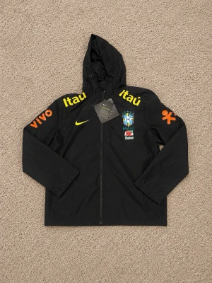 Svart Nike Brasil jacka med huva - Snygg svart träningsjacka från Nike med huva och dragkedja. Jackan har Brasilien fotbollslandslagets logga på bröstet, gula och orangea sponsorloggor samt Nike Swoosh. Perfekt för dig som gillar sportig stil och vill visa stöd för Brasilien.