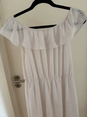 Vit offshoulder sommarklänning - Söt vit långklänning med offshoulder-modell och volang upptill. Klänningen har broderade hål och resår i midjan för en bekväm passform. Perfekt för varma dagar och har en romantisk känsla.
