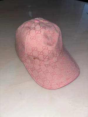 Rosa keps från Gucci - Snygg rosa keps från Gucci med klassiskt GG-monogram över hela kepsen. Kepsen har böjd skärm och är tillverkad i canvasmaterial. Perfekt accessoar för att lyfta din streetwear-look.