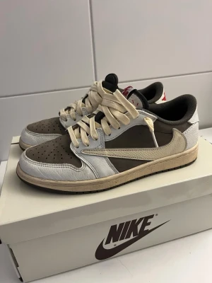 Nike Air Jordan 1 Low Travis Scott - Nike Air Jordan 1 Low Travis Scott i brunt, vitt och beige läder med ikonisk omvänd Swoosh och röda broderade detaljer på hälen. Skorna har svarta och gråa inslag samt tjocka beige skosnören. Perfekt för dig som gillar streetwear och unika sneakers.