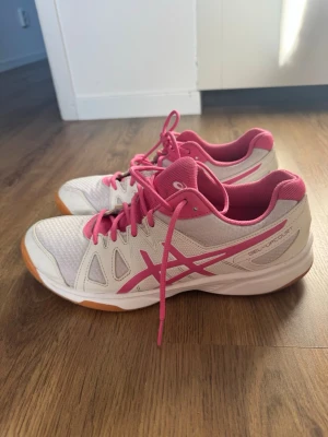 Vita och rosa Asics inomhusskor - Säljer ett par vita Asics GEL-UPCOURT inomhusskor med rosa detaljer och snörning. Skorna har en slitstark gummisula i orange, ventilerande mesh och syntetiska förstärkningar. Perfekta för sporter som badminton eller volleyboll. priset kan diskuteras