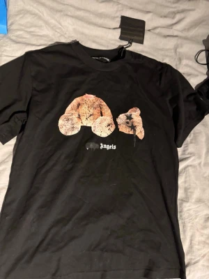 Svart Palm Angels t-shirt med nalletryck - Cool svart t-shirt från Palm Angels med ett unikt tryck av en nallebjörn på bröstet och vit logga under. Klassisk passform och rund halsringning. Perfekt för dig som gillar streetwear och vill sticka ut med en statement-tee.