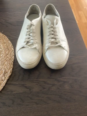 Vita sneakers från Axel Arigato - Stilrena vita sneakers från Axel Arigato i lågt snitt. Skorna har minimalistisk design med vita skosnören och diskret logga på hälen. Tillverkade i slätt läder med klassisk rund tå och tjock vit sula. Perfekta för en clean och modern look.
