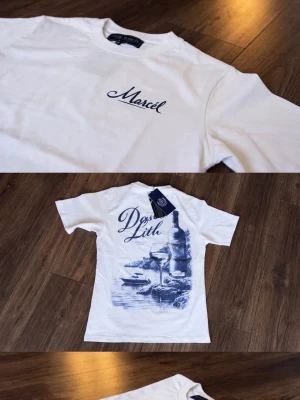 Vit t-shirt från Leon Marcel  - Vit t-shirt från Leon Marcel  med blå tryckdetaljer. Framsidan har texten 'Marcel' och baksidan har ett stort motiv med vinflaska, glas och havsmotiv i blått. Klassisk passform och rund halsringning. Perfekt för en avslappnad stil.