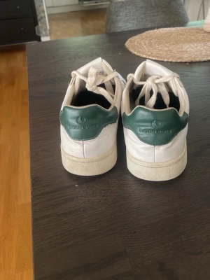 Vita Björn Borg sneakers med gröna detaljer - Snygga vita sneakers från Björn Borg med gröna detaljer på hälen och klassisk låg profil. Skorna har vita snören och är tillverkade i slätt syntetmaterial med diskret logga på baksidan och plösen. Perfekta för en clean och stilren look.