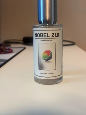 NOBEL 21.0 parfym från One Bold Chemist - Fräsch parfym i glasflaska med metallock från One Bold Chemist, modell NOBEL 21.0. Flaskan är genomskinlig med en etikett som har en färgglad citrusfrukt på framsidan. Parfymen har en ljusgul nyans och modern design.