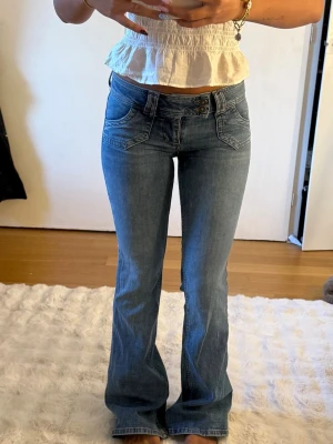 Blå bootcut jeans med låg midja - Säljer ett par snygga blå bootcut jeans med låg midja och dubbla knappar framtill. Jeansen har klassiska fickor både fram och bak och är tillverkade i ett mjukt denimtyg.
