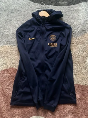 Mörkblå PSG träningsjacka Nike - Snygg mörkblå träningsjacka från Nike med Paris Saint-Germain-logga och Qatar Airways-tryck i guld. Jackan har hel dragkedja, huva och långa ärmar. Perfekt för fotbollsträning eller chill. Materialet är troligtvis polyester för sportig känsla.