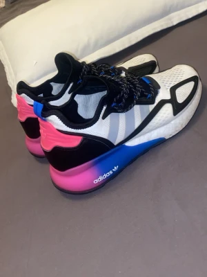 Adidas sneakers i vitt, svart och rosa - Snygga Adidas sneakers med vit mesh-överdel, svarta detaljer och rosa samt blå färgskiftning på sulan. Skorna har svarta och vita snören, samt en cool design med 3 ränder på sidan och rosa hälparti. Perfekta för dig som gillar färgstarka och moderna sneakers.