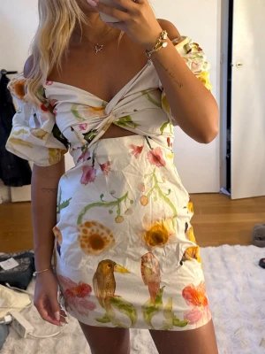 Vit miniklänning med blommmönster - Superfin vit miniklänning med färgglada blommor och fåglar, puffärmar och twistad detalj framtill. Klänningen har off-shoulder design och är gjord i ett lätt tyg, perfekt för sommaren och Midsommar