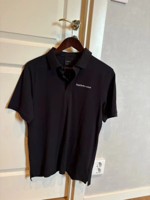 Ny piké från Peak Performance - Svart polotopp från Peak Performance med klassisk krage och korta ärmar. Diskret logotyp broderad på bröstet. Toppens material är mjuk bomull och passformen är normal, perfekt för en clean och stilren look.