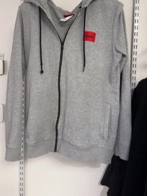 Grå hoodie med dragkedja från HUGO - Snygg grå hoodie från HUGO med svart dragkedja och svarta snören i huvan. På bröstet sitter en röd patch med HUGO-loggan. Hoodien har fickor på sidorna och är tillverkad i mjukt bomullsmaterial. Perfekt för en chill och stilren look.