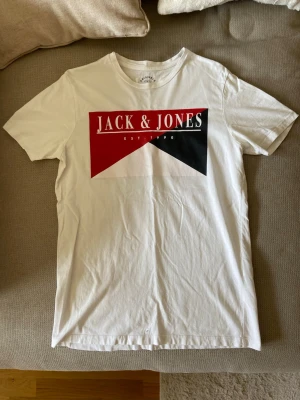 Vit Jack & Jones t-shirt med tryck - Snygg vit t-shirt från Jack & Jones med stort grafiskt tryck i rött, svart och vitt på bröstet. Klassisk rund hals och korta ärmar. Perfekt till jeans eller shorts för en avslappnad look.