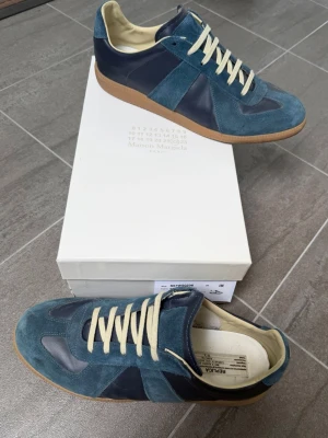 Blå sneakers Maison Margiela - Snygga sneakers från Maison Margiela i en mix av mörkblått läder och blå mocka. Skorna har beige innersula, vita snören och klassisk gummisula. Perfekt för dig som gillar stilrena och exklusiva detaljer.