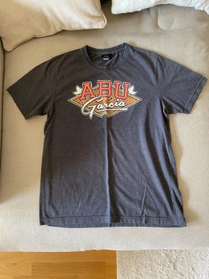Mörkgrå ABU Garcia t-shirt  - Snygg mörkgrå t-shirt från South West med stort ABU Garcia-tryck i rött, vitt och gult på bröstet. Klassisk passform och rund hals. Perfekt för dig som gillar streetwear och vill ha en unik look.