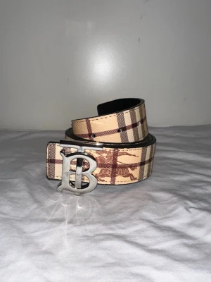 Beige rutigt Burberry bälte - Snyggt bälte från Burberry i beige och brunt med klassiskt rutigt mönster och silverfärgat B-spänne. Bältet har detaljer med Burberrys ikoniska hästmotiv och är tillverkat i skinn. Perfekt accessoar för att lyfta din outfit.