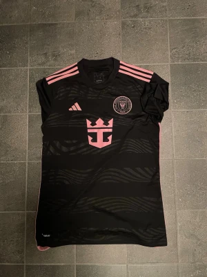 Inter Miami Messi matchtröja svart/rosa - Snygg svart Inter Miami matchtröja från Adidas med rosa detaljer och Messi 10 på ryggen. Tröjan har klubbmärke på bröstet, rosa ränder på axlarna och är tillverkad i ett lätt, ventilerande material perfekt för fotboll eller träning.
