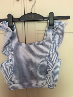 Randig blåvit linnetopp med volang - Superfin croppad linnetopp från Zara i blåvitt randigt med volangdetaljer längs axelbanden. Toppen har fyrkantig ringning och knäpps med en knapp i ryggen. Perfekt för varma dagar och ger en fräsch sommarkänsla.