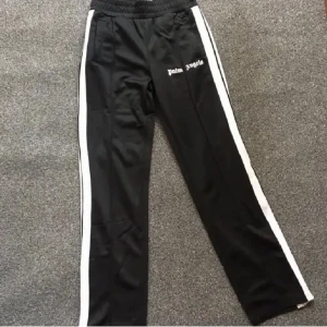 Palm Angels svarta trackpants  - Svarta trackpants från Palm Angels med vita sidoränder och broderad logga på ena benet. Byxorna har elastisk midja med snörning och är tillverkade i 100% polyester för en sportig vibe. Perfekta för en streetwear-look.