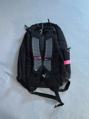 Svart Nike Elite ryggsäck med rosa detaljer - Snygg svart Nike Elite ryggsäck med rosa logga och detaljer. Ryggsäcken har vadderade axelremmar, flera fack och meshficka på insidan. Perfekt för skolan eller träningen, med sportig design och slitstarkt material.