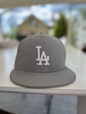 Grå LA-keps från New Era - Snygg grå keps från New Era med LA-logga broderad i vitt framtill. Klassisk flat brim-modell och tillverkad i polyester. Perfekt för dig som gillar streetstyle och sportiga vibes.