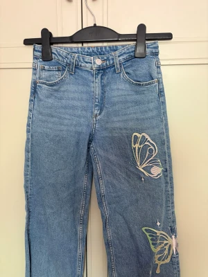 Blå jeans med broderade fjärilar - Snygga blå jeans med raka ben och broderade fjärilar i pastellfärger på ena benet. Klassisk femficksmodell med knapp och dragkedja. Low waist