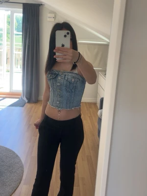 Denim korsett-topp med knappar - Snygg croppad korsett-topp i ljusblå denim med knappar framtill och smockad rygg. Toppen är ärmlös och har en cool, edgy vibe som passar perfekt till både jeans och kjol. Perfekt för dig som vill sticka ut med en trendig look.