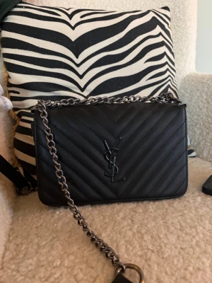 Svart axelväska från Saint Laurent - Snygg svart axelväska från Saint Laurent i quiltat läder med kedjerem i silverfärg. Väskan har YSL-logga framtill och stilrena, diagonala sömmar som ger en lyxig känsla. Perfekt storlek för det viktigaste och en tidlös design som lyfter varje outfit.