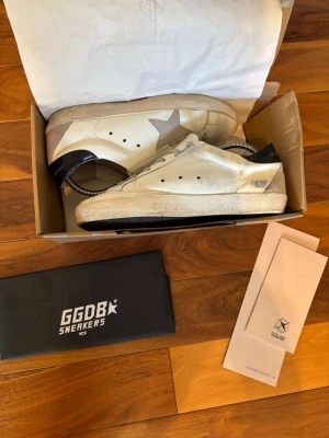 Golden Goose vita sneakers med stjärna - Snygga vita sneakers från Golden Goose med grå stjärndetalj på sidan och svart hälparti. Skorna har klassisk låg profil, vita snören och slitstark gummisula. Perfekta för dig som gillar en trendig och avslappnad stil. Storlek 42! Skriv om ni har några funderingar!
