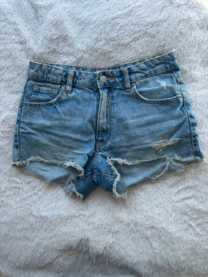 Blå jeansshorts från Gina Tricot - Säljer ett par blå jeansshorts från Gina Tricot med råa kanter och slitna detaljer. Perfekta för sommaren och ger en avslappnad vibe.