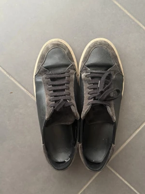 Axel Arigato bruna sneakers – Premium lädersneakers | Mycket fint skick - Säljer ett par svarta sneakers från Axel Arigato med ovandel i läder och mocka. Skorna har svarta snören, låg profil och vit sula. Märket syns diskret i guld på sidan. Perfekta för en clean och stilren look.