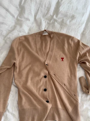 Beige kofta från Ami Paris - Säljer en beige kofta från Ami Paris med svart knappstängning och det ikoniska röda Ami de Coeur-logotypen broderad på bröstet. Koftan har ribbade muddar och är tillverkad i mjukt stickat material. Perfekt för lager på lager och stilrena outfits.