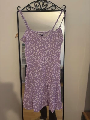 Ljuslila blommig kortklänning - Söt ljuslila kortklänning med vita små blommor och smala axelband. Klänningen har snörning i ryggen för en justerbar passform och är tillverkad i ett lätt och luftigt material, perfekt för varma dagar.