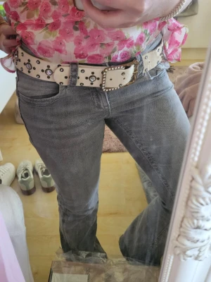 Vitt bälte med nitar och strassdetaljer - Snyggt vitt bälte med glittrande strass runt det fyrkantiga spännet och dekorativa nitar längs hela bältet. Bältet har en cool och trendig look som passar perfekt till jeans eller kjol. Jag har fått ärva detta bälte av min mamma så jag vet inte märket. Bältet är använt men i bra skick. 