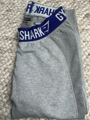 Grå Gymshark tights med blå resår - Snygga grå tights från Gymshark med bred blå resår i midjan och vit logga. Tillverkade i ett stretchigt och snabbtorkande material som passar perfekt till träning. Tightsen har en tight passform och är designade för att sitta bekvämt under hela passet.