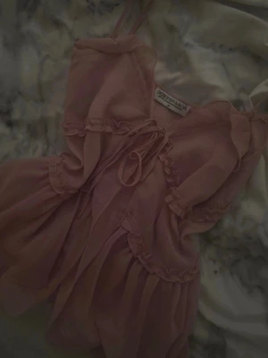Rosa volangblus från Designbyamor - Supersöt rosa blus från Designbyamor med tunna axelband och volangdetaljer. Blusen har knytning framtill och är gjord i ett lätt, transparent tyg som ger en romantisk vibe. Perfekt för dig som gillar en feminin och trendig stil.