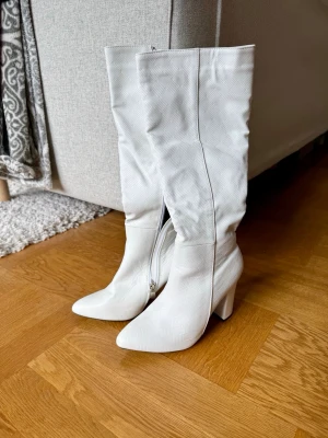 Vita höga boots med klack - Säljer ett par vita höga stövlar med spetsig tå och bred klack. Stövlarna har dragkedja på insidan och ett diskret rutmönster i materialet. Finns två märken, kolla tredje bilden