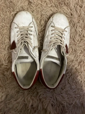 Philippe Model vita sneakers - Snygga vita sneakers från Philippe Model med röda detaljer och grå inslag på sulan. Skorna har en klassisk låg siluett, vita skosnören och en ikonisk sköldlogga på sidan. Materialet är en mix av läder och textil för en modern look.